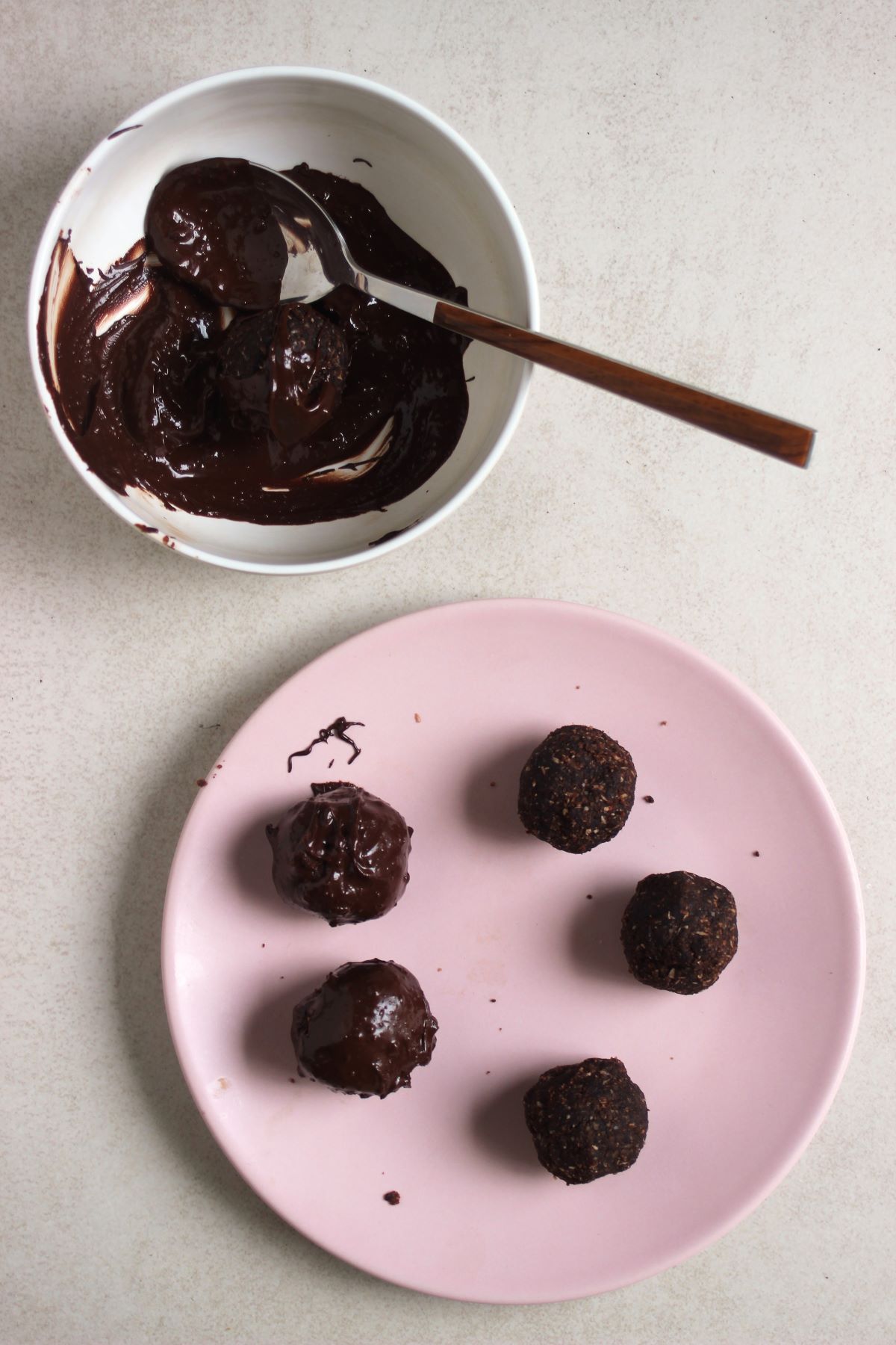 Chocolate peanut butter balls on a pink plate, two with chocolate coating. Another bowl with melted chocolate and a spoon.
