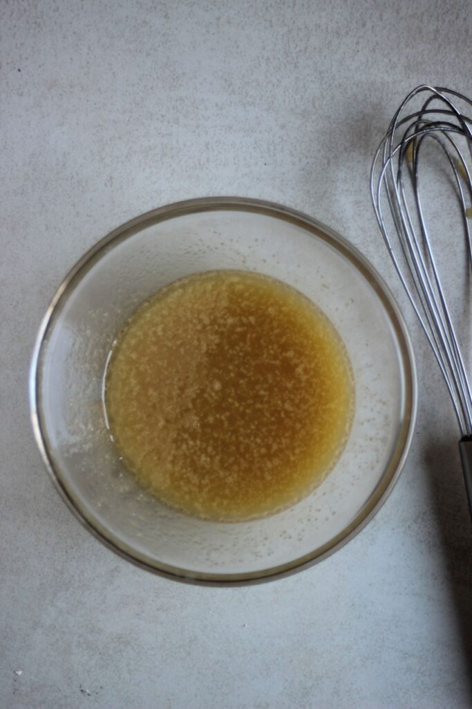 Glass bowl with a brown/yellow liquid. A hand whisk on the side.