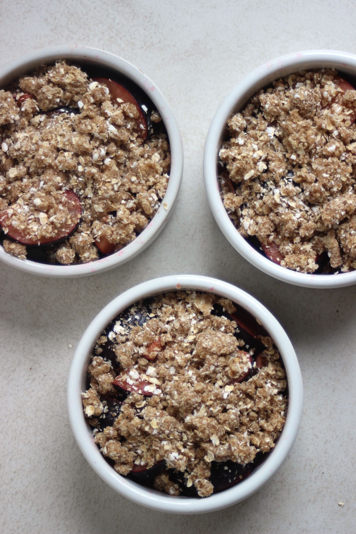 Three ramekins with plum crisp before bake on a white surface seen from above.
