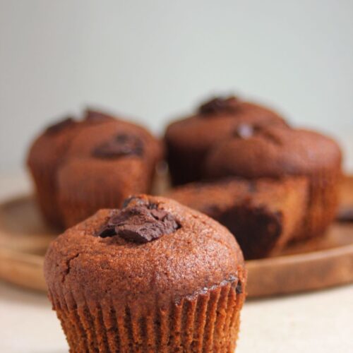 A chocolate banana muffin, and more muffins behind on a wooden plate.