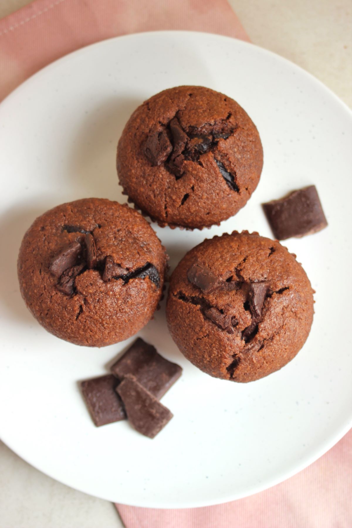 Three banana chocolate muffins on a white plate seen from above. Chocolate chunks on the side.
