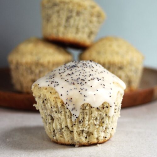 A lemon muffin with poppy seeds on a white surface. More muffins behind.