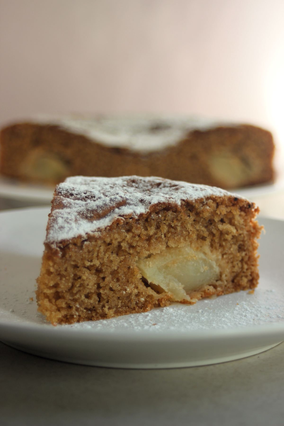A portion of apple cake sprinkle with powdered sugar on a white plate. The cake behind.