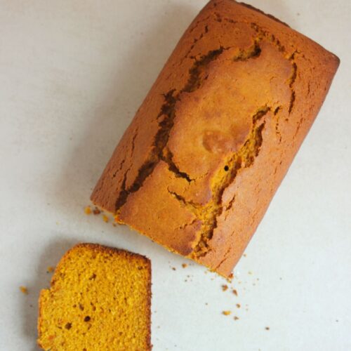 A pumpkin bread and a slice of it on a white surface seen from above.