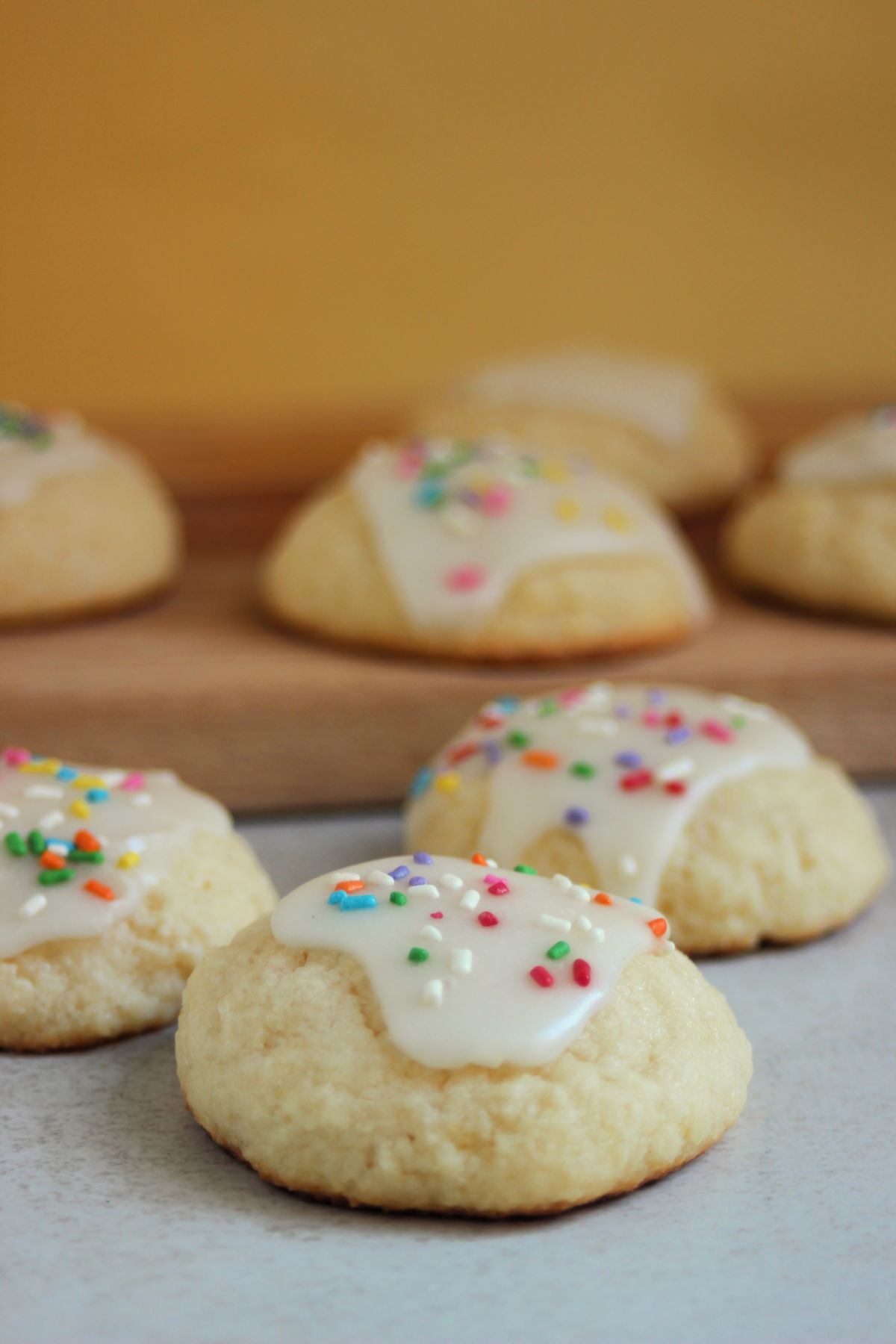 Ricotta cookies with icing and sprinkles. More cookies behind on a wooden board.