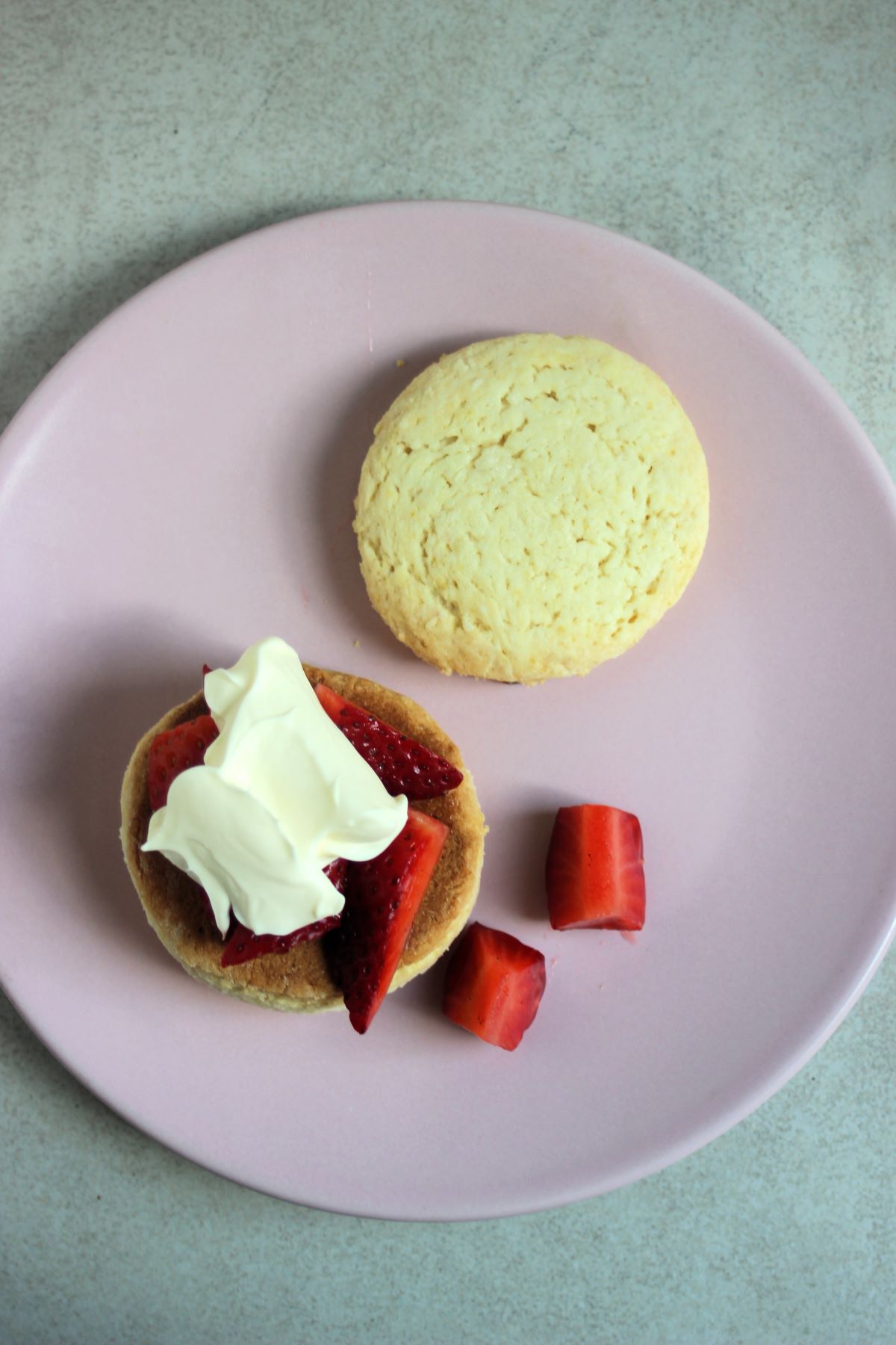 Shortcake assembly. Two biscuits of a shortcake on a pink plate, one with strawberries and cream.