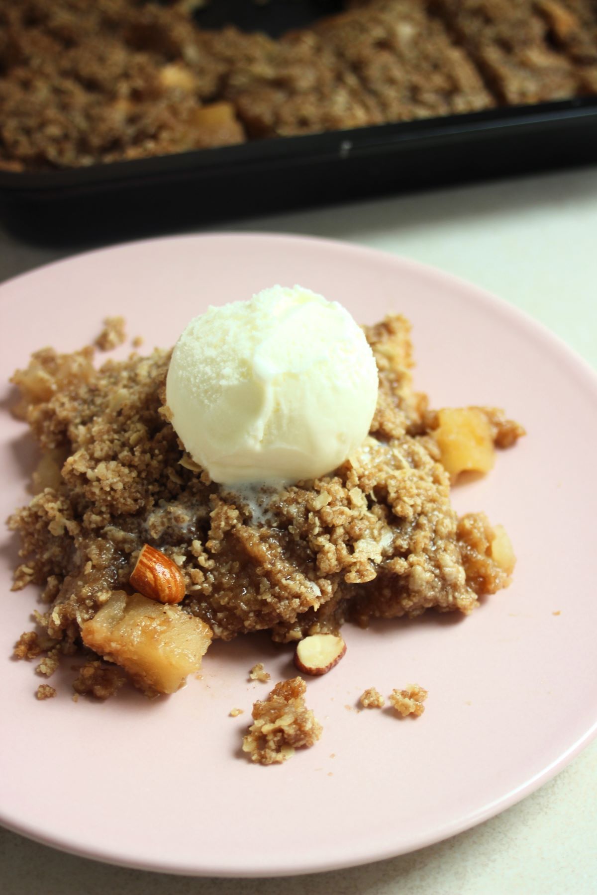 A portion of apple crisp on a pink plate with a scoop of ice cream above.