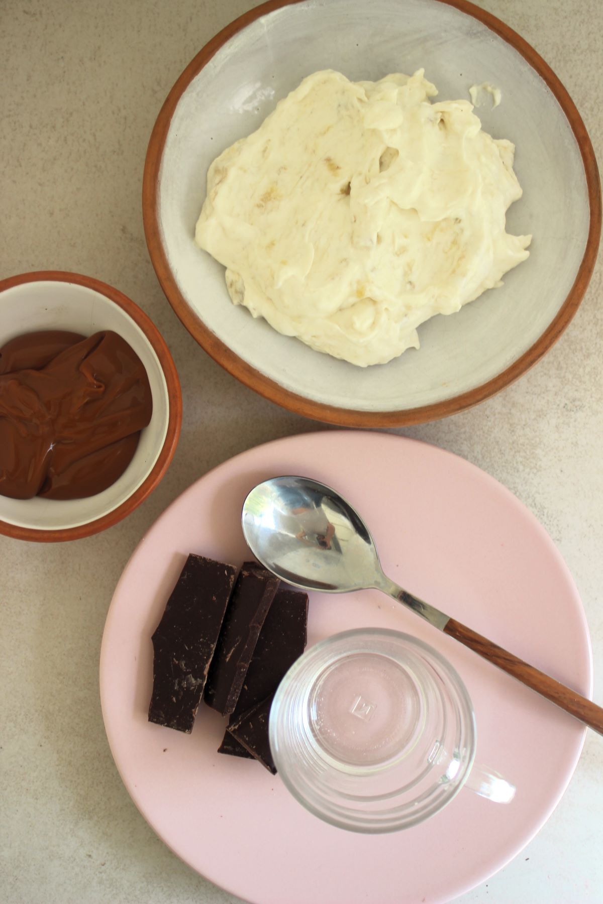 Bowl with dulce de leche, a plate with banana cream, and another plate with chocolate, a glass, and a spoon, seen from above.