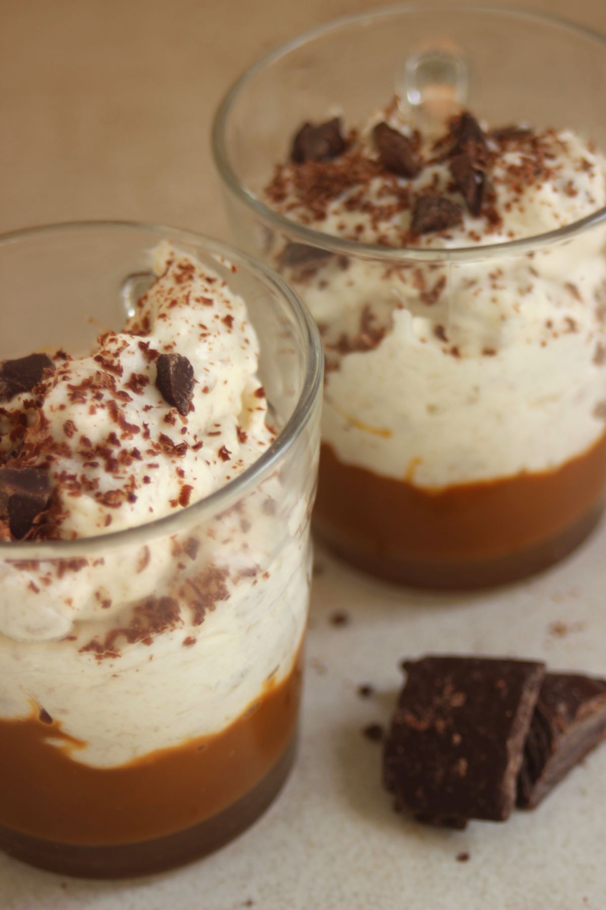 Two glasses with dulce de leche, banana cream, and shredded chocolate. Chocolate chunks on the surface.