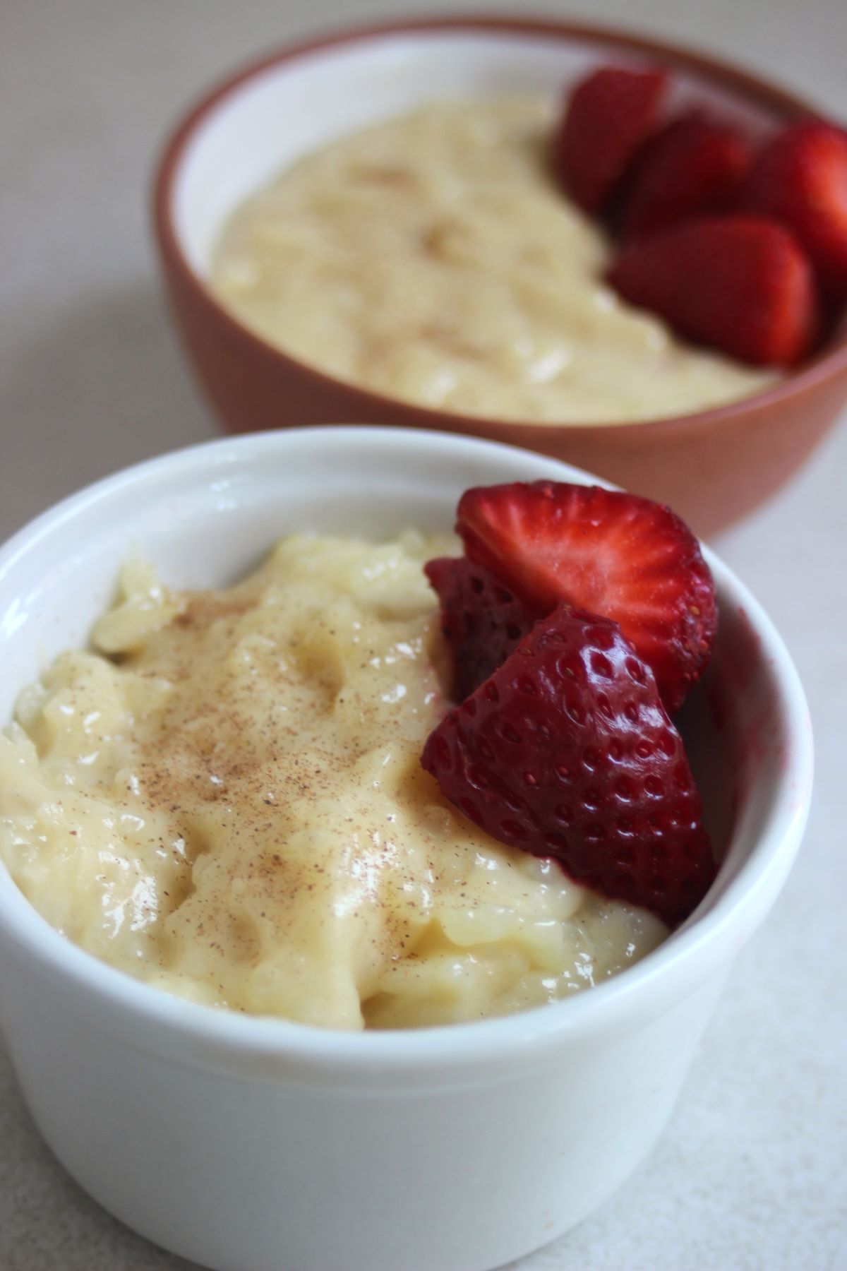 White ramekin with caramel rice pudding and fresh strawberries. Another bowl with rice pudding behind.