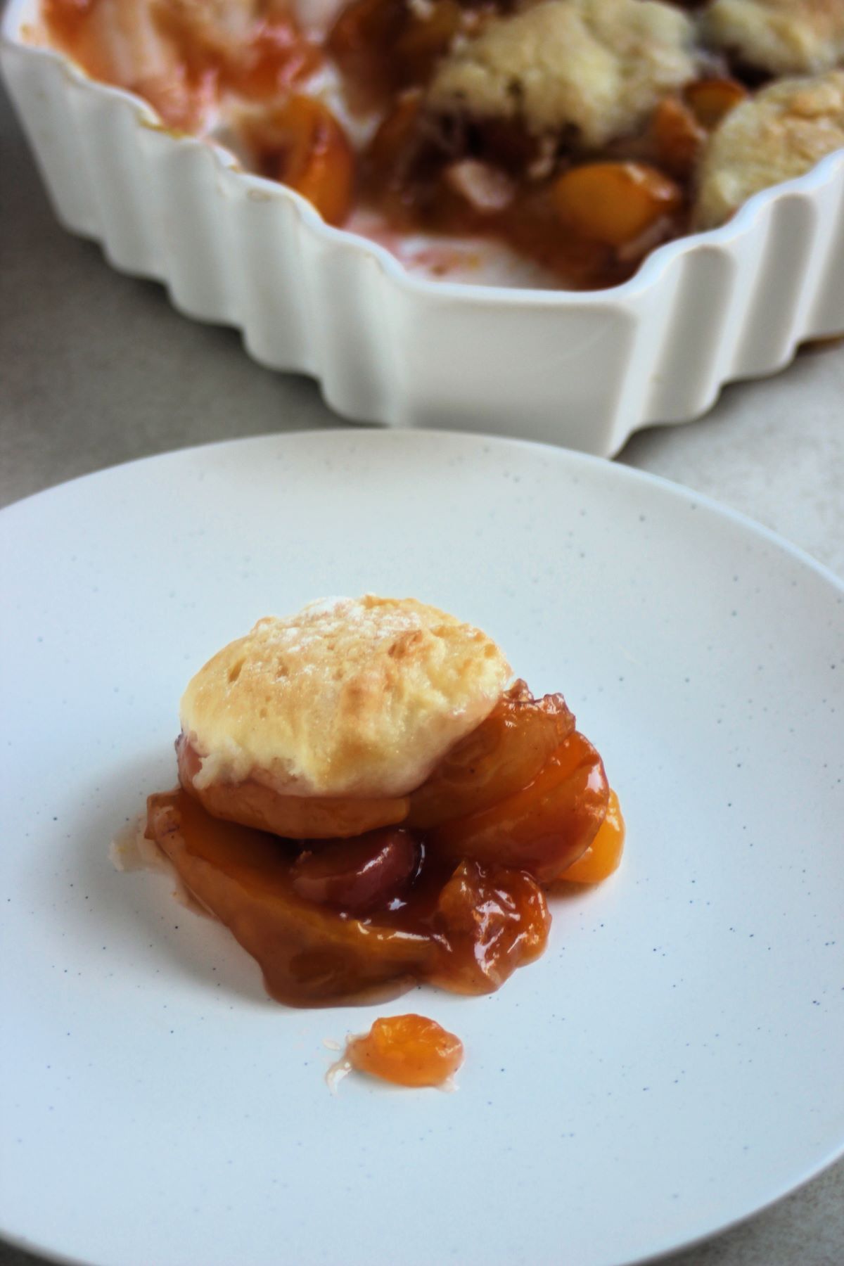 Portion of peach cobbler on a white plate and a baking dish with peach cobbler behind.