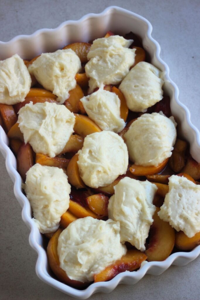 Rectangular baking dish with sliced peaches and scoops of batter.