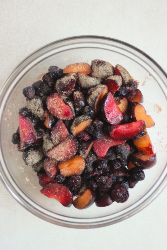 Glass bowl with slice of plums, mixed berries, and sugar.