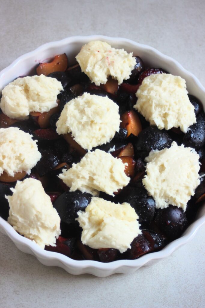 Round baking pan with plums, mixed berries, and spoons of batter on top.