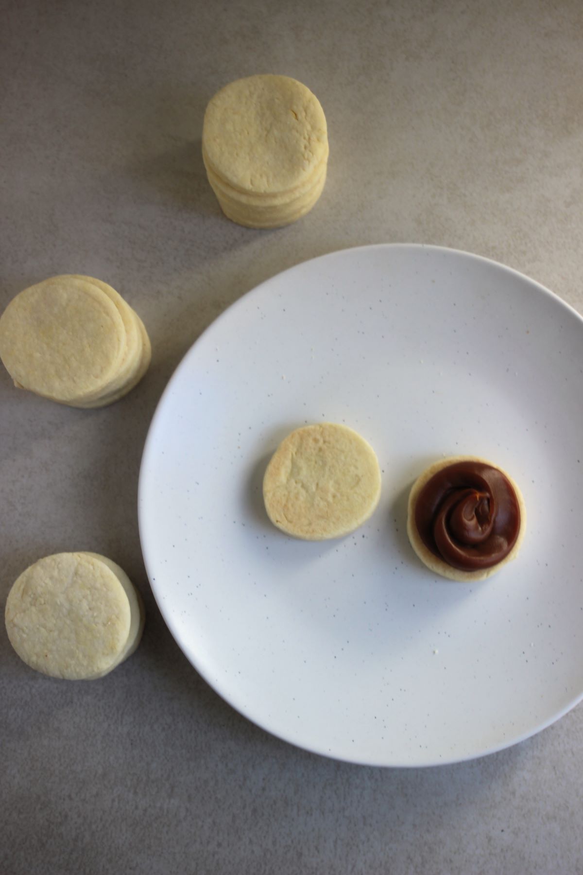 Two circles of alfajor dough, one with dulce de leche, on a white plate. Many round circles of alfajor dough on the side.