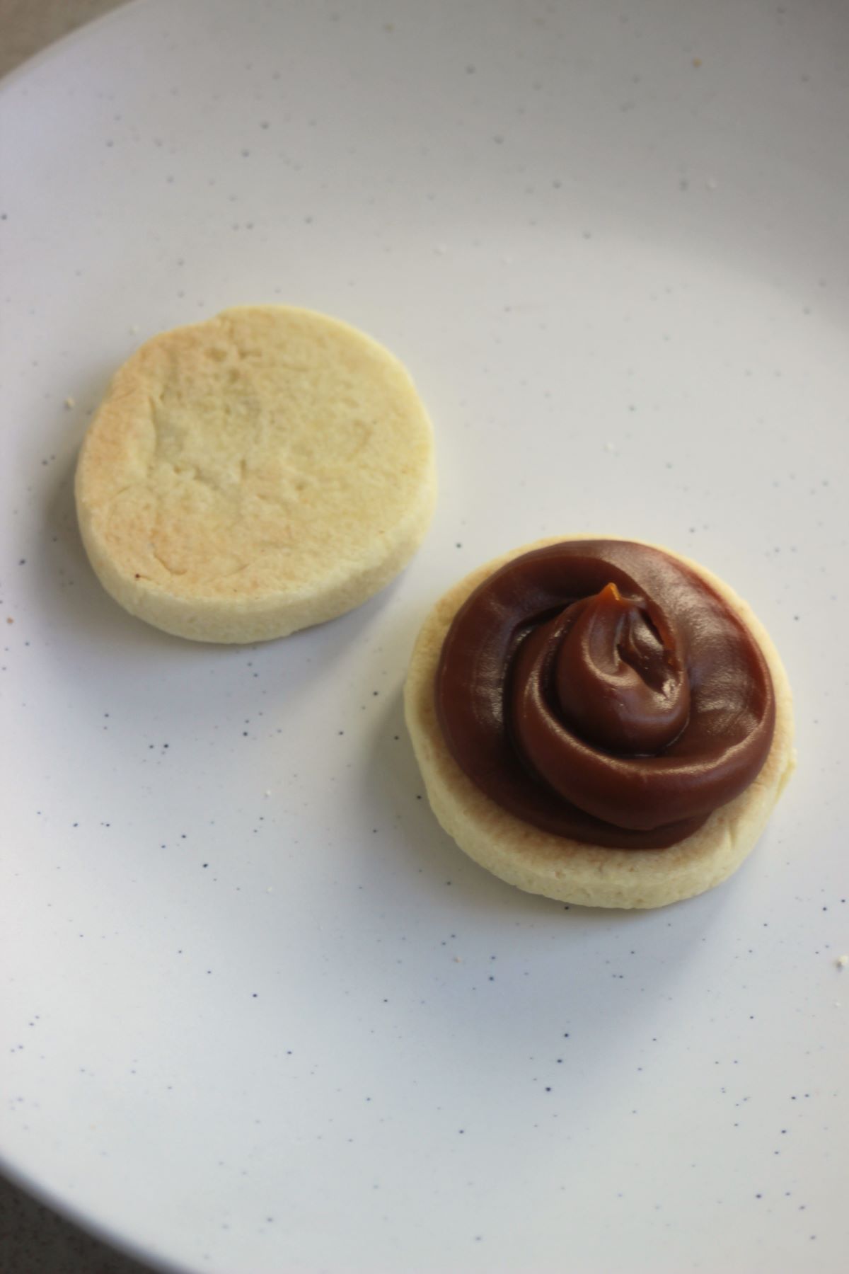 Two circles of alfajor dough, one with dulce de leche, on a white plate.