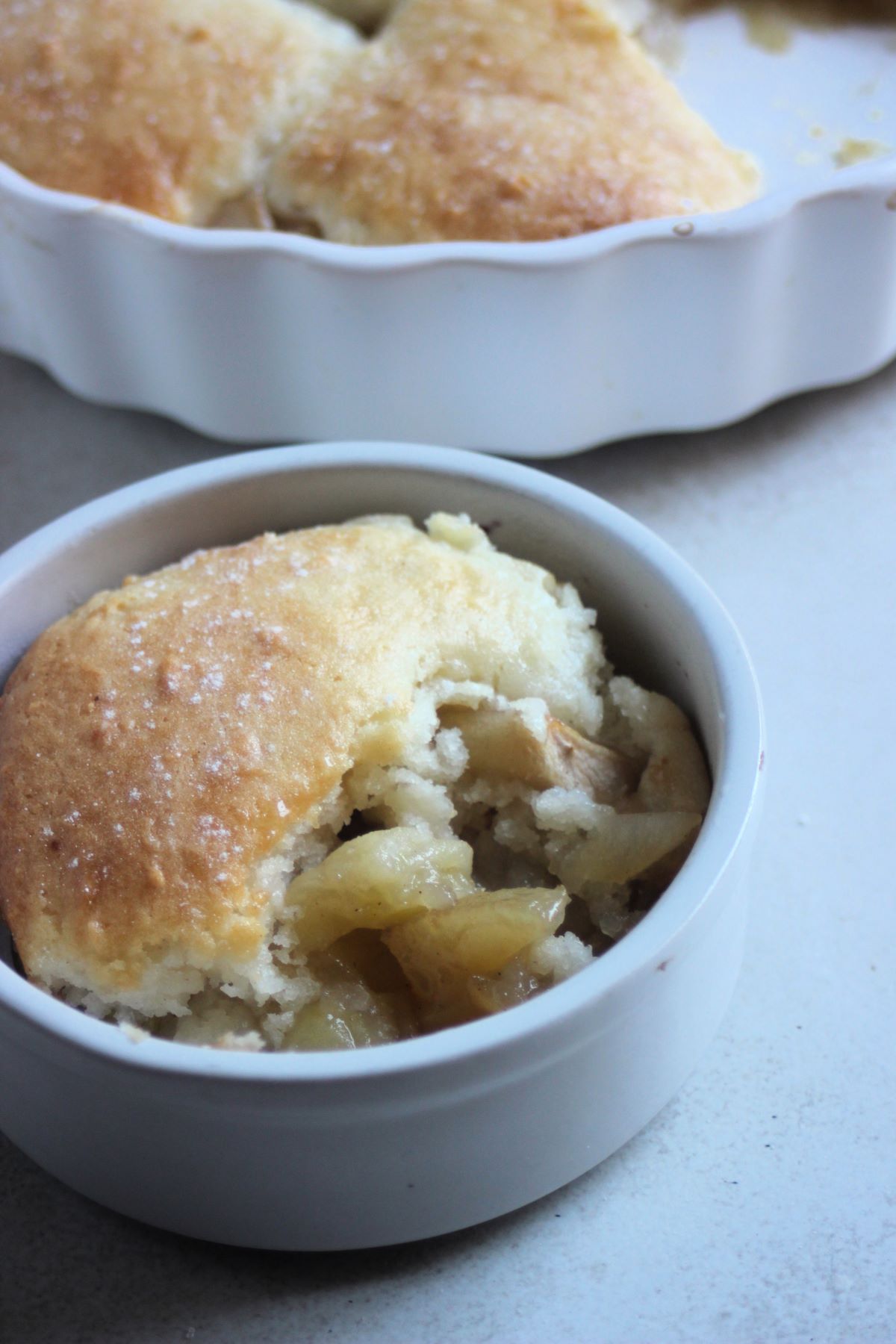 Small bowl with a portion of apple cobbler. Apple cobbler behind.