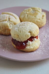 Scon filled with cream cheese and jam on a pink plate. More scons behind.