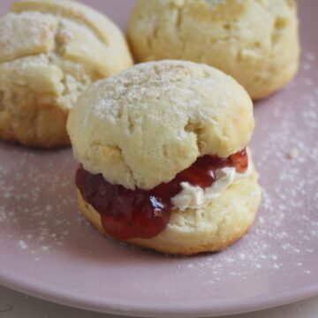 Scon filled with cream cheese and jam on a pink plate. More scons behind.