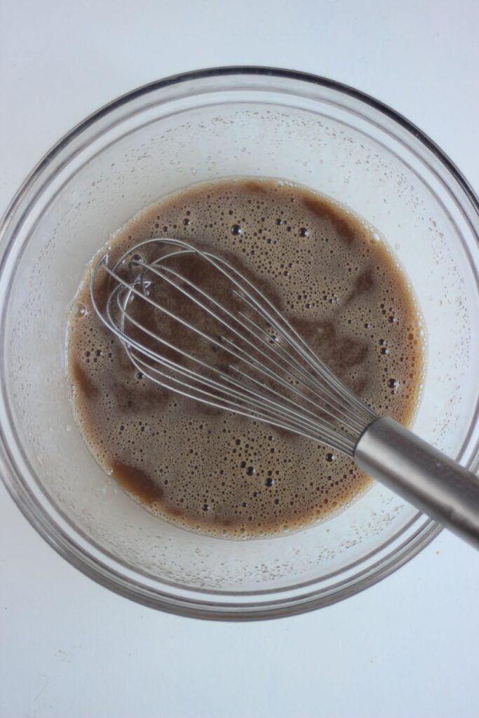 Glass bowl with a brown liquid and a hand whisk.