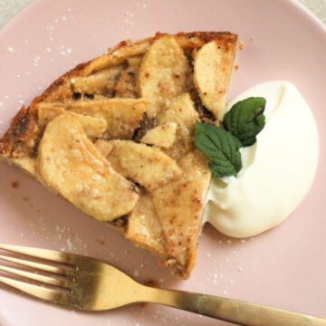 Portion of apple cake with whipped cream and a golden fork on a pink plate.