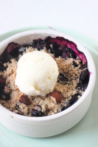 Blueberry crisp with a scoop of vanilla ice cream in a white ramekin on a aqua green plate.