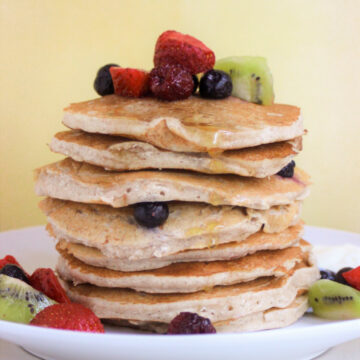 A tower of pancakes with strawberries, blueberries, and kiwis on a white plate. Yellow background.