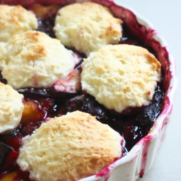 Round white baking dish with plum cobbler on a white surface.