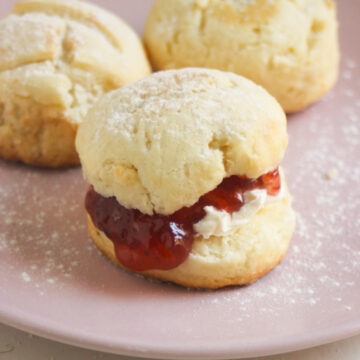 Scon filled with cream cheese and jam on a pink plate. More scones behind.