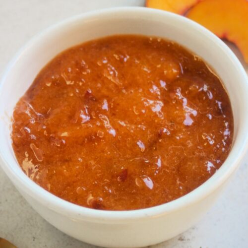 White small bowl with peach jam on a white surface. Golden spoon and fresh peach slices on the sides.