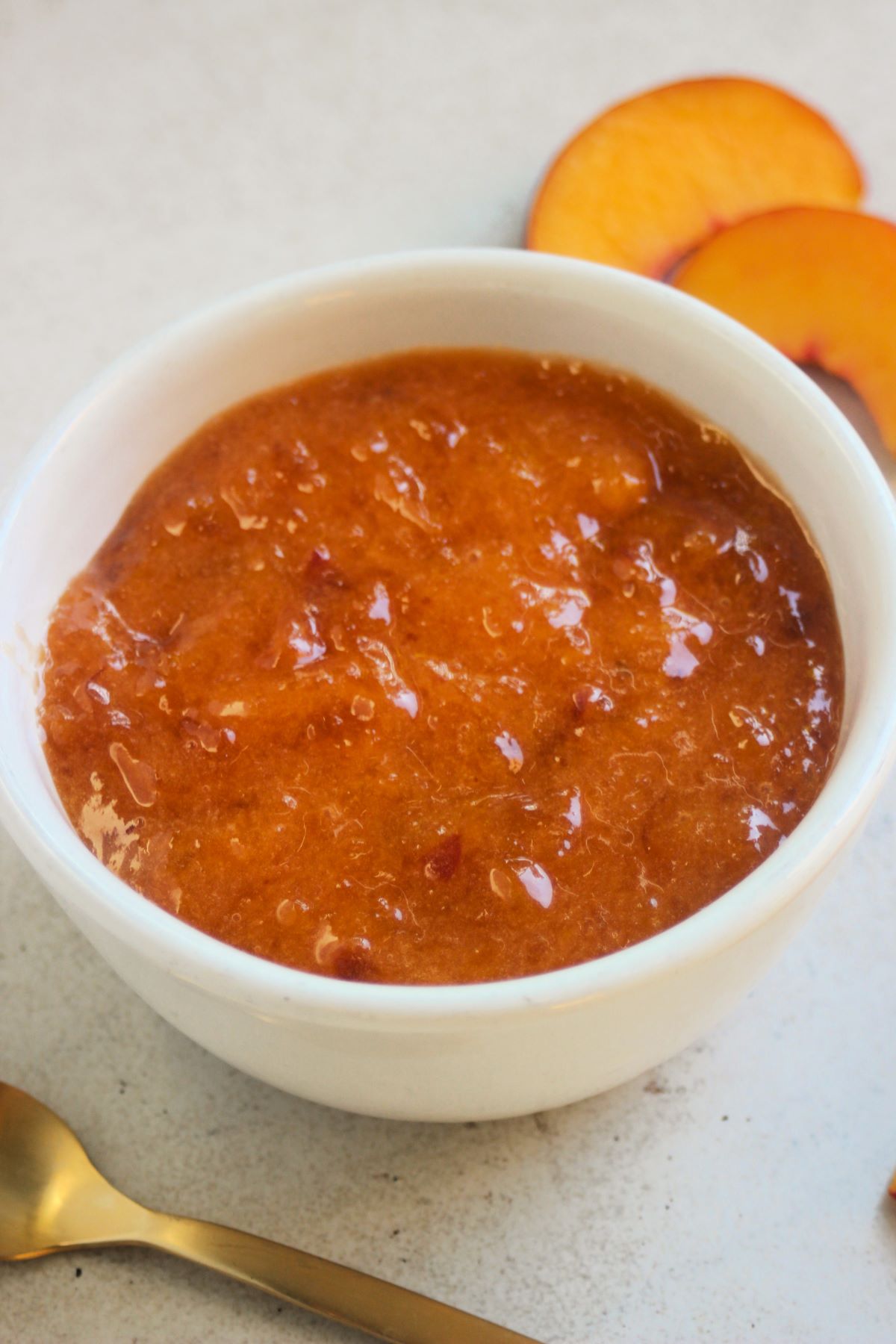 White small bowl with peach jam on a white surface. Golden spoon and fresh peach slices on the sides.