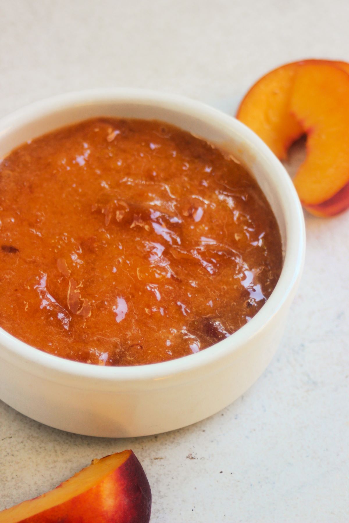 White small bowl with peach jam on a white surface. Fresh peach slices on the sides.