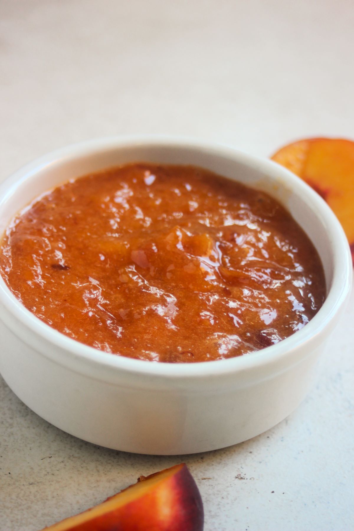 White small bowl with peach jam on a white surface. Fresh peach slices on the sides.