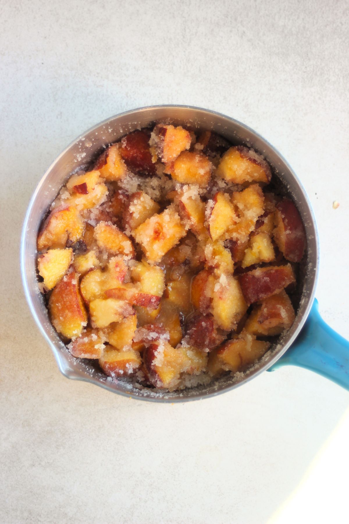 Deep pan with chopped peaches and sugar on a white surface seen from above.