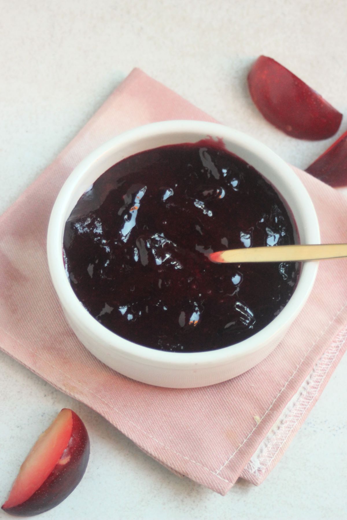 Plum jam on a small white round bowl and golden spoon. Pink napkin below. Fresh plum slices on the sides.