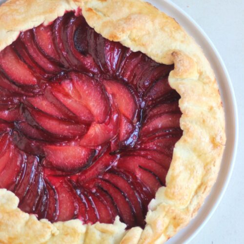 Plum tart on a white plate on a white surface seen from above.