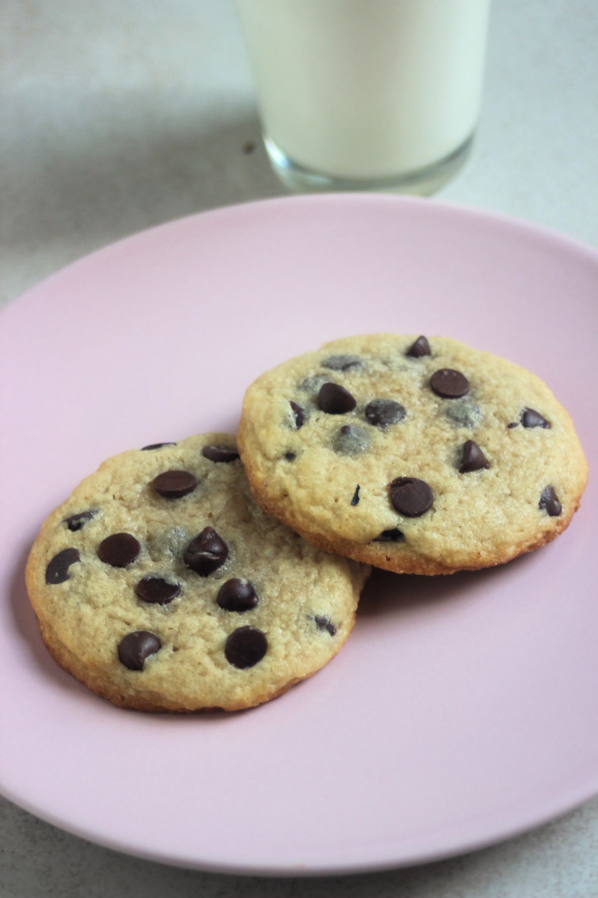 Two chocolate chip cookies on a pink plate. The bottom of a glass of milk behind.