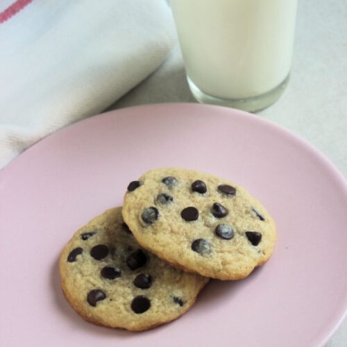 Two chocolate chip cookies on a pink plate. A glass of milk and a white napkin behind.