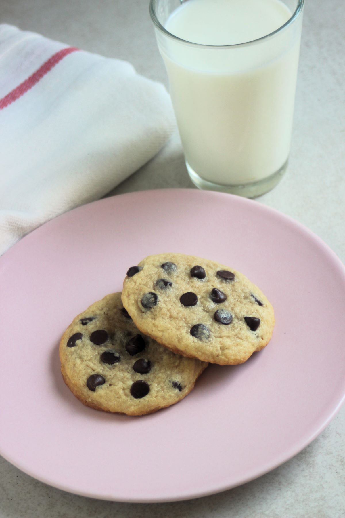 Two chocolate chip cookies on a pink plate. A glass of milk and a white napkin behind.