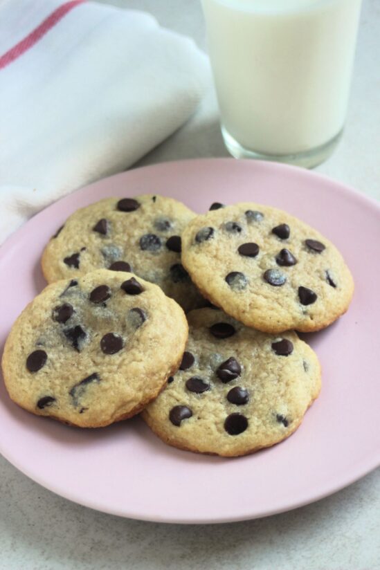 Four chocolate chip cookies on a pink plate. A glass of milk and a white napkin behind.