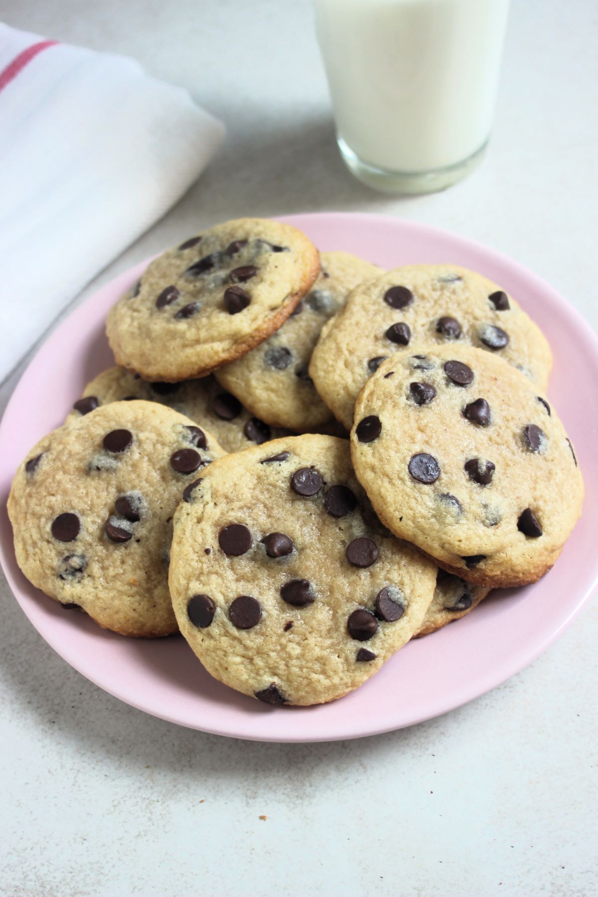 Many chocolate chip cookies on a pink plate, A glass of milk, and a white napkin behind.