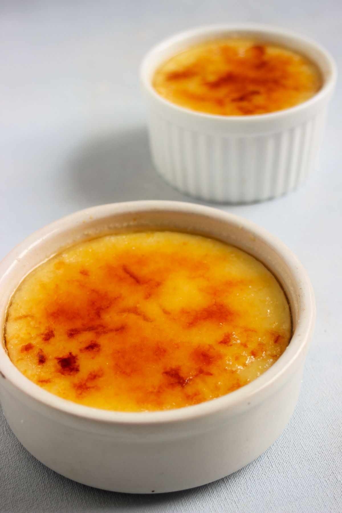 White ramekin with crema catalana and behind another small ramekin with crema catalana on a light blue surface.