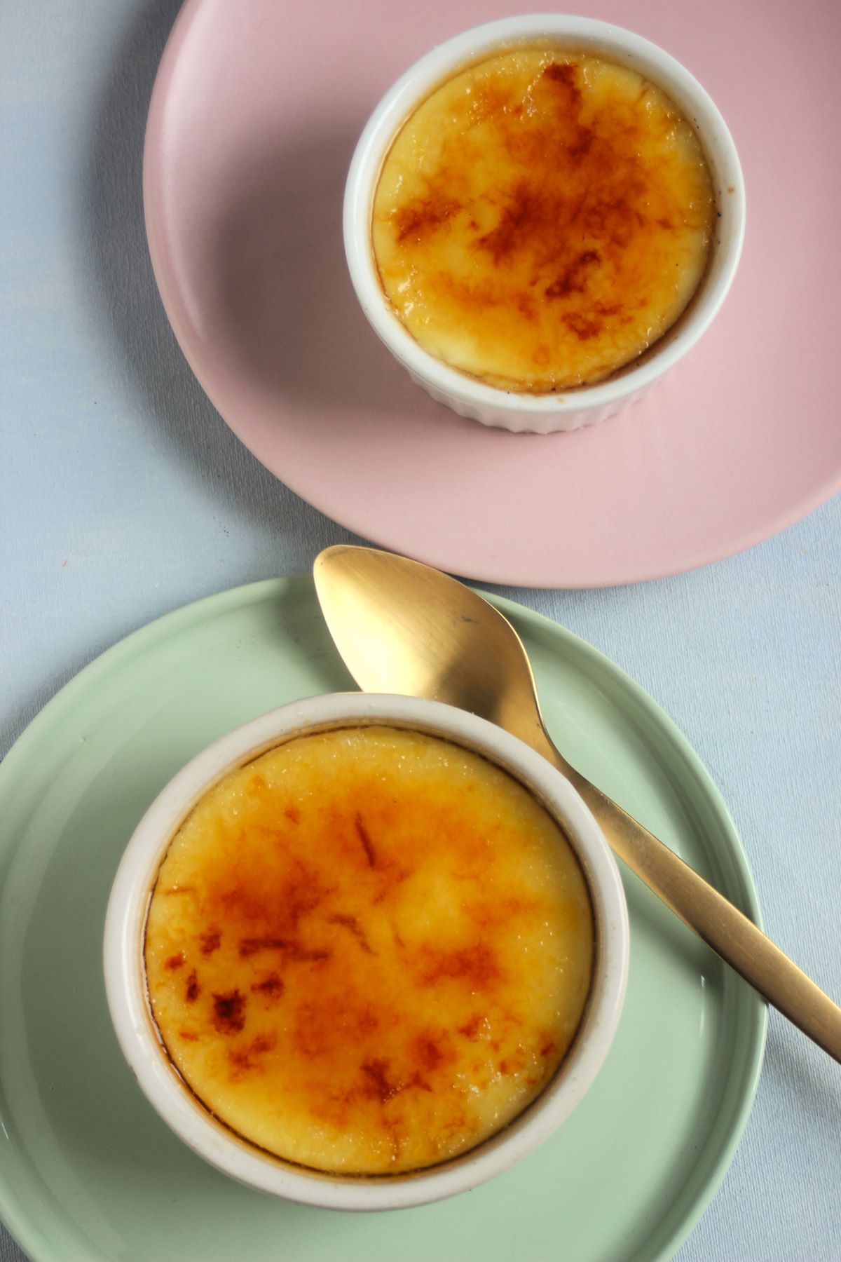 Two ramekins with crema catalana seen from above. One in a pink plate and the other in an aqua-green plate with a golden fork.