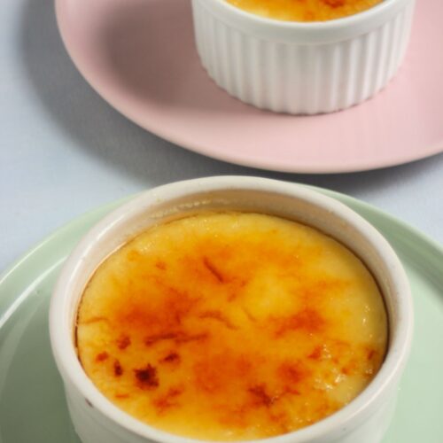 Ramekin with crema catalana on an aqua-green plate and behind a small ramekin with crema catalana on a pink plate.