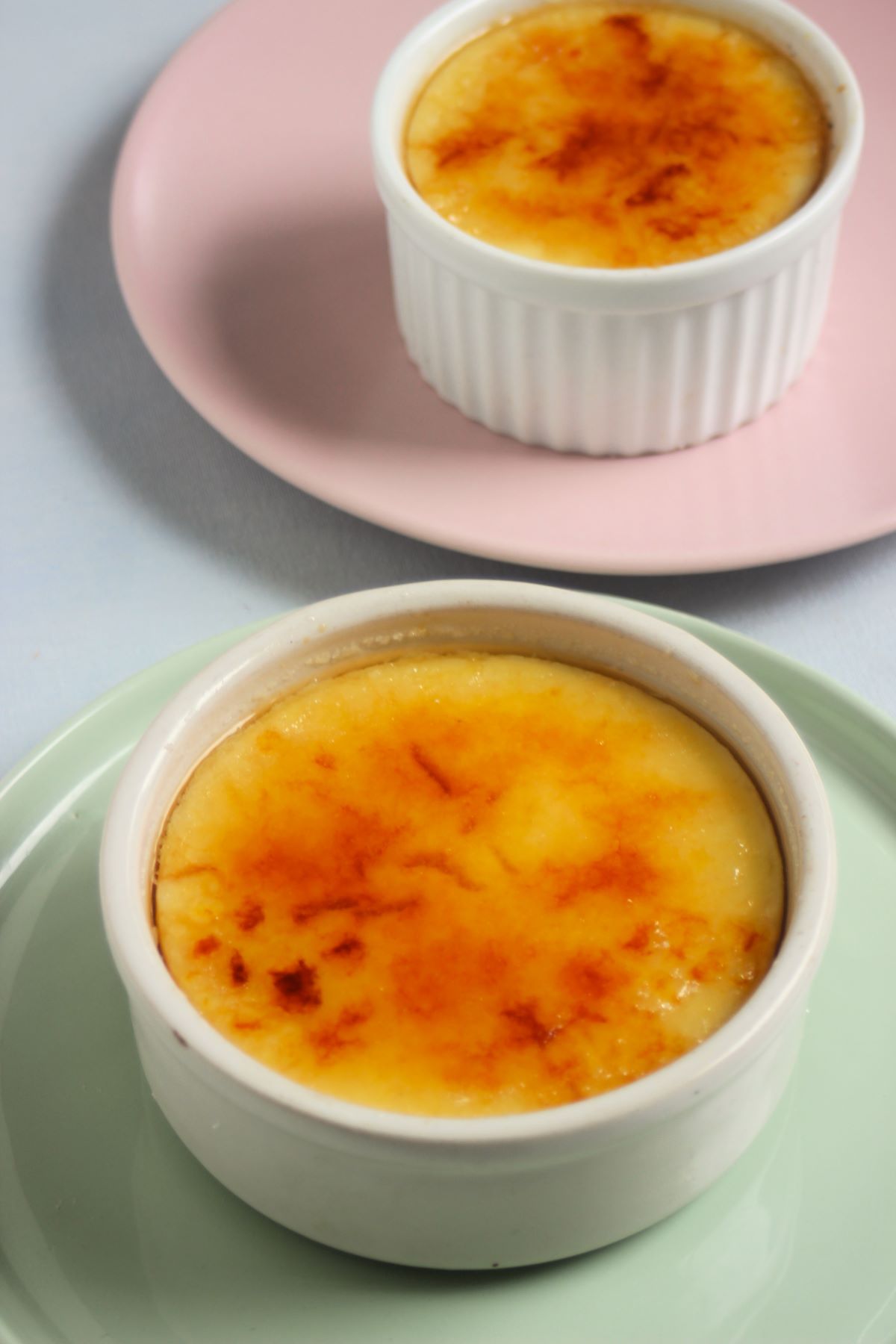 Ramekin with crema catalana on an aqua-green plate and behind a small ramekin with crema catalana on a pink plate.