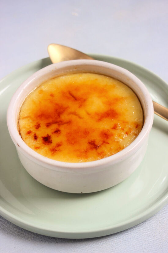 White ramekin with crema catalana on an aqua-green plate and a golden spoon.