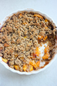 White round baking dish with peach crisp with a quarter eaten, on a white surface seen from above.