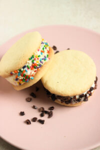 Two ice cream cookie sandwich , one leaning on the other, and cocoa nibs on a pink plate.