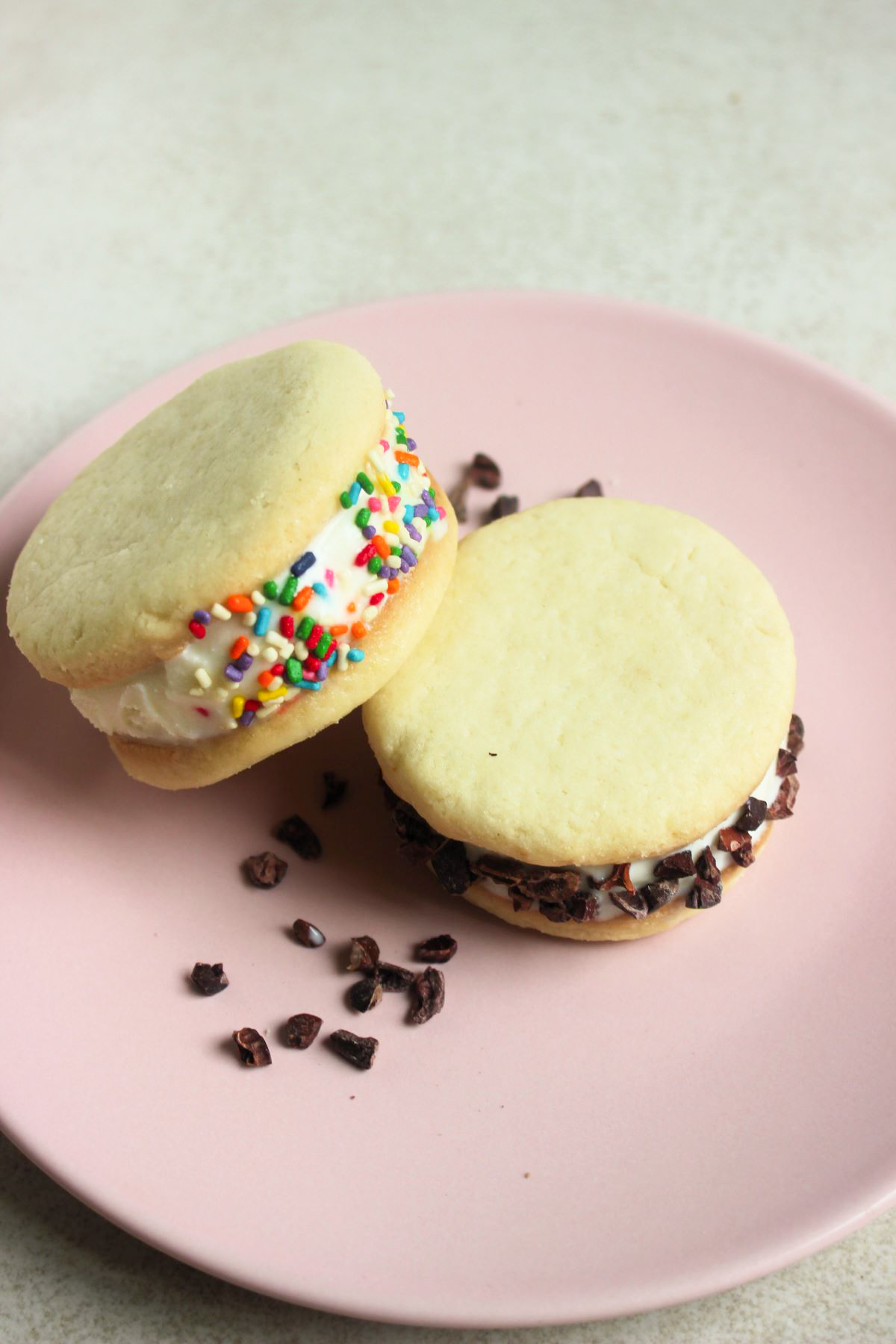 Two ice cream cookie sandwich , one leaning on the other, and cocoa nibs on a pink plate.