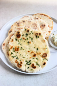 Naan breads on a white plate. The first one is brush with herb butter.